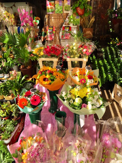A vibrant display of fresh floral bouquets arranged on a pink table in a floral shop. The bouquets feature a variety of colorful flowers including bright pink gerbera daisies, yellow sunflowers, white