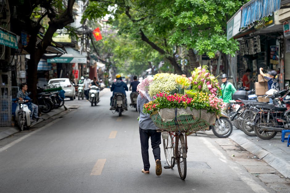 A street scene on Upton Park High Street amidst a vibrant market atmosphere, featuring a person pushing a bicycle laden with a large, colorful floral arrangement. The bouquet, arranged with a variety