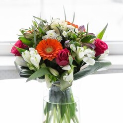 This striking floral arrangement, meticulously assembled by seasoned Upton Park florists, offers an exuberant yet refined display of colour and composition. At its centre, four large gerbera daisies stand out-two glowing with fiery orange vibrancy, two radiating in creamy peach, each crowned with a contrasting brown centre. Bold roses in shades of magenta and fuchsia nestle between, their tightly coiled petals adding an element of romance familiar to those who wander the shops near the Boleyn Ground. The arrangement sparkles with sprays of white alstroemeria, their delicate, multi-petaled blooms contributing textural interest alongside petite white and pale pink flowers peeking from the abundance of greenery. Surrounding the bloom cluster, broad, paddle-shaped aspidistra leaves are wrapped in elegant curves, providing a lush, verdant base that anchors the design and recalls the green heart of Central Park. Tall, clear glass contains the bouquet, revealing a dynamic tangle of fresh green stems that promise longevity and vitality. Soft daylight, reminiscent of sunlight filtering through an East London bay window, bathes the entire composition in clarity, highlighting the interplay of light, shadow, and hue. This arrangement balances joy, celebration, and sophistication, perfect for gifting on any occasion from new beginnings to heartfelt thanks, infusing each home in Upton Park with its abundant spirit and professional finesse.